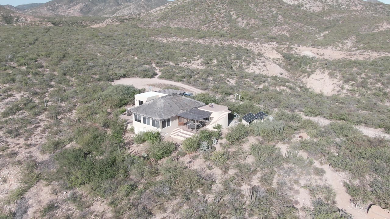 Aerial view of a light-colored house in Cabo Pulmo, Baja California Sur, surrounded by desert scrub, cacti, dirt tracks, and rocky hills, with no visible people.