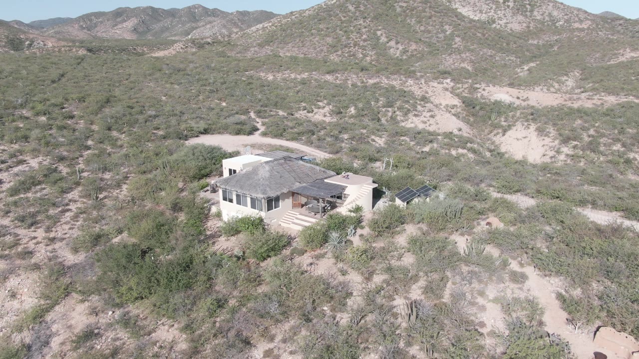 Aerial frame of the Cabo Pulmo desert house with pale walls, a dark roof, nearby solar panels, scrub vegetation, sandy tracks, and distant hills, with no visible people.