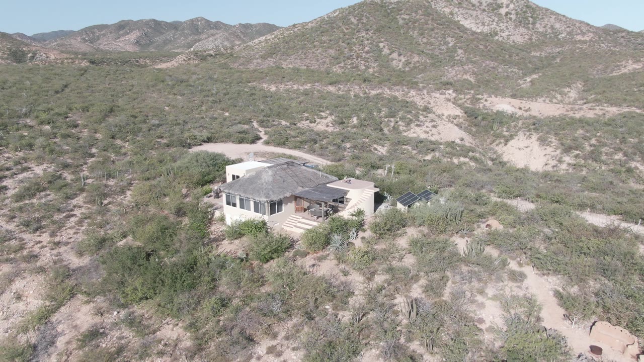 Wide aerial view in Cabo Pulmo showing a solitary desert house below low rocky mountains, with cactus-dotted scrub, a dirt road, solar panels, and no visible people.