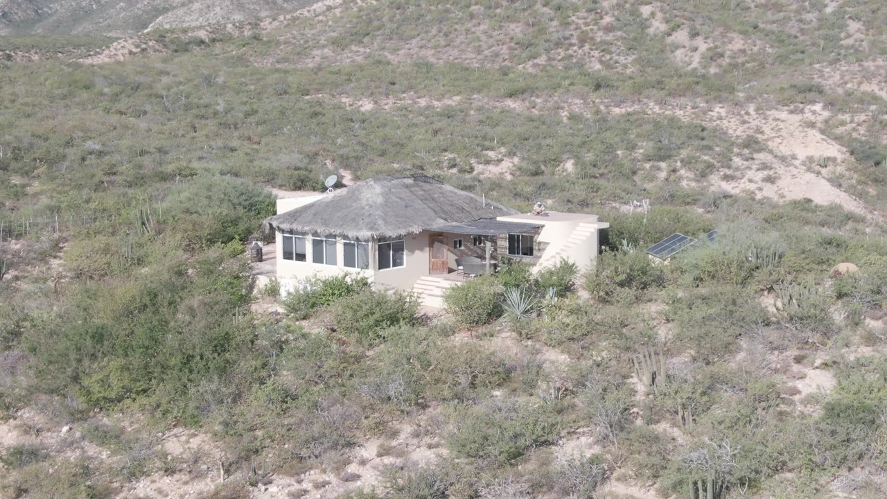 A light-colored single-story house in Cabo Pulmo, Baja California Sur sits in arid shrubland with cacti and low hills behind it, with no visible people.