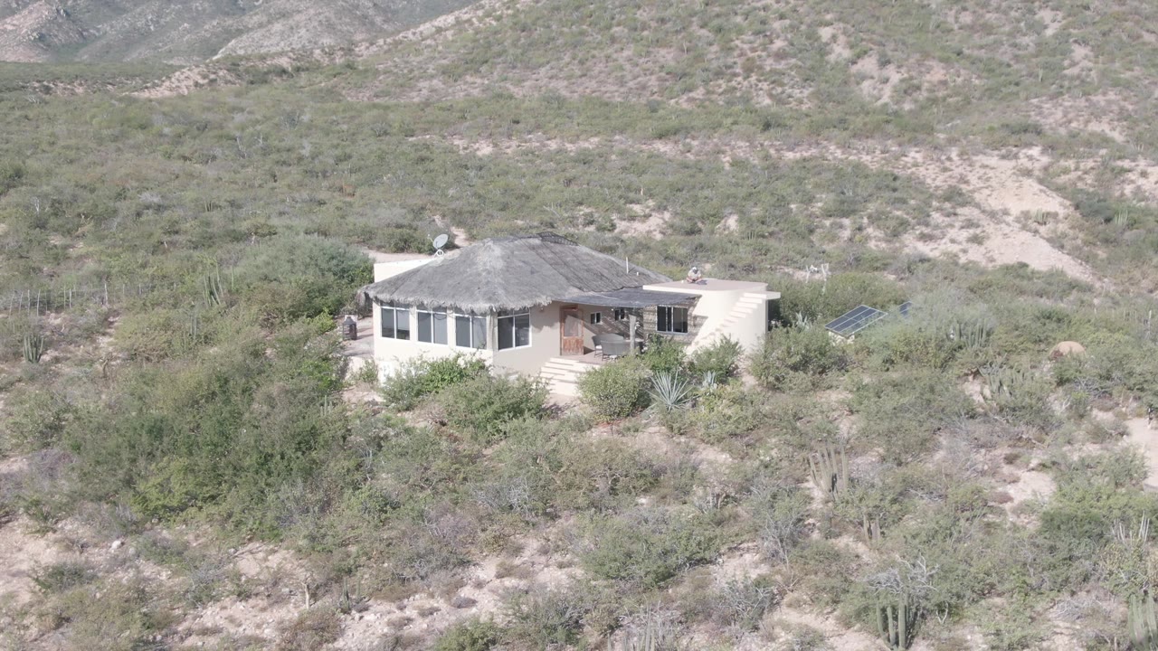 The Cabo Pulmo desert house remains centered in the frame, surrounded by scrubby vegetation, cacti, sandy washes, and muted rocky hills, with no visible people.