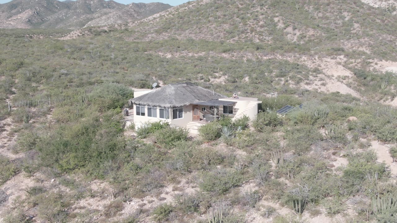 A single-story house in Cabo Pulmo, Baja California Sur appears slightly right of center amid pale scrub, cacti, and dry hillside terrain, with no visible people.