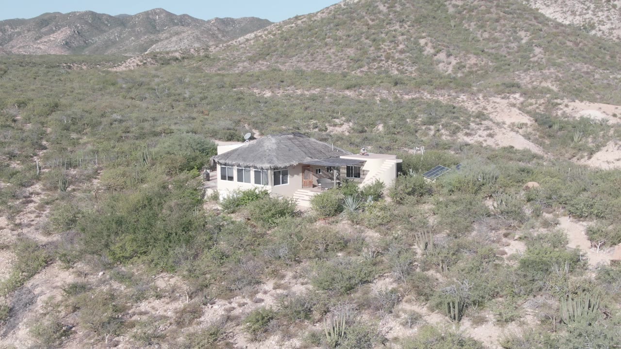 Remote desert house with thick foreground brush and low mountains behind.