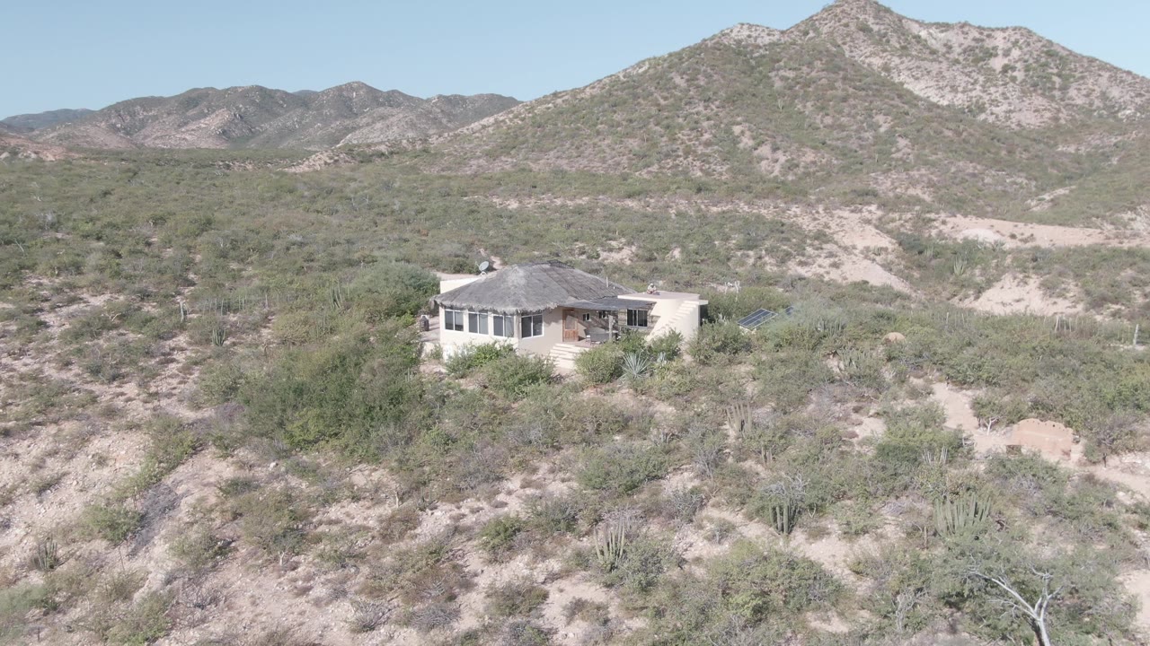 A light-colored house sits in Cabo Pulmo desert brush below rocky mountains, with sandy washes, scattered cacti, and no visible people at the start of the clip.