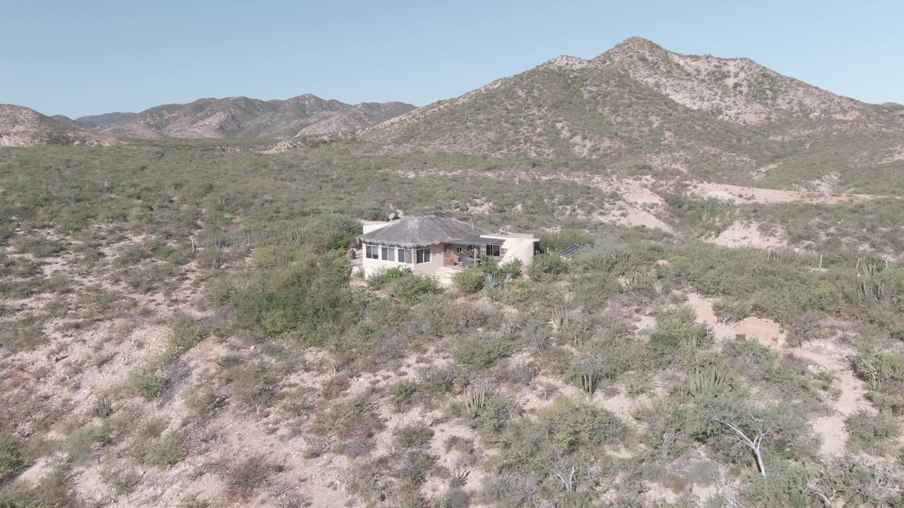 A high view shows the small Cabo Pulmo house partly surrounded by green-brown desert shrubs, with pale sandy ground and layered mountains beyond, no visible people.