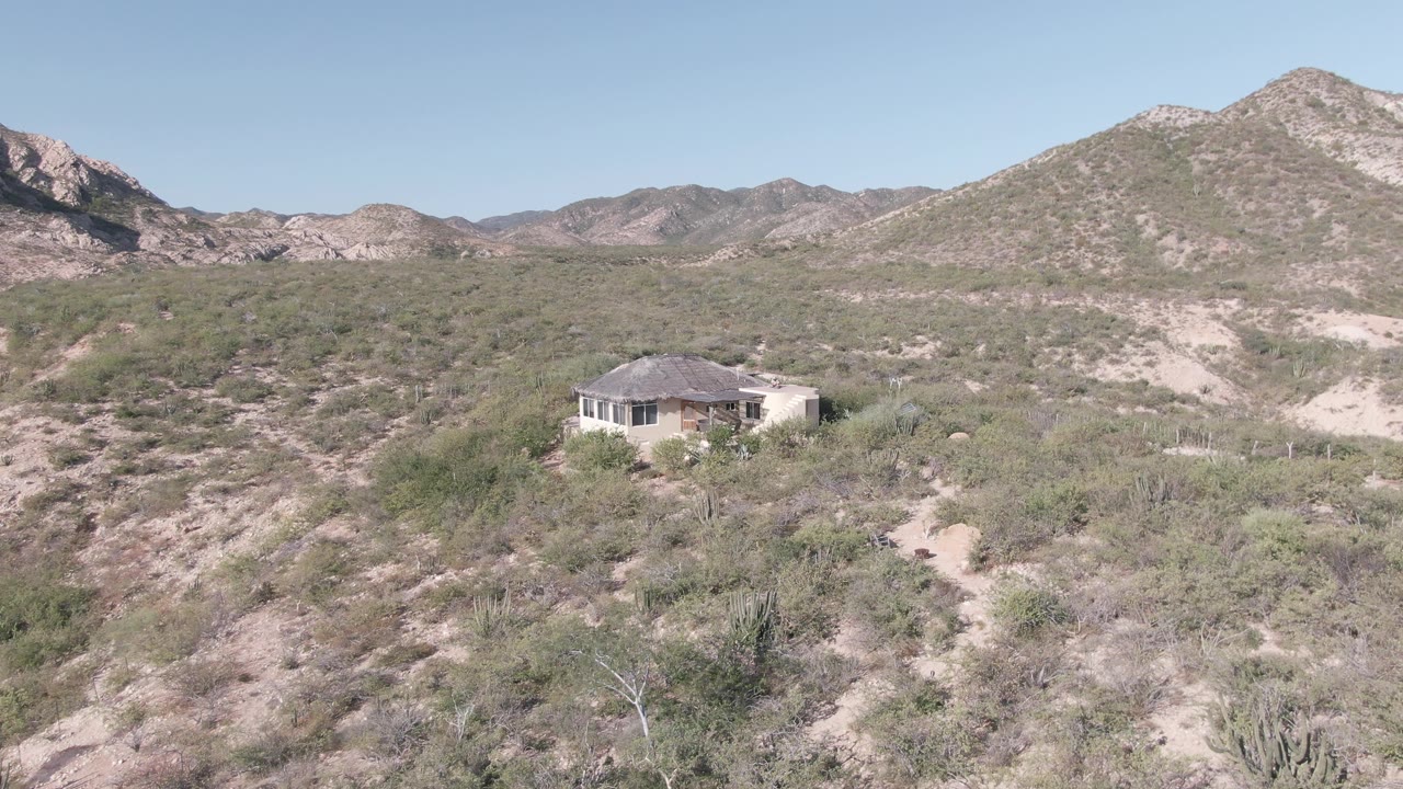 Wide elevated view of a small house in the Cabo Pulmo desert, with sandy ground, sparse shrubs, cactus-like plants, pale mountain ridges, clear sky, and no visible people.