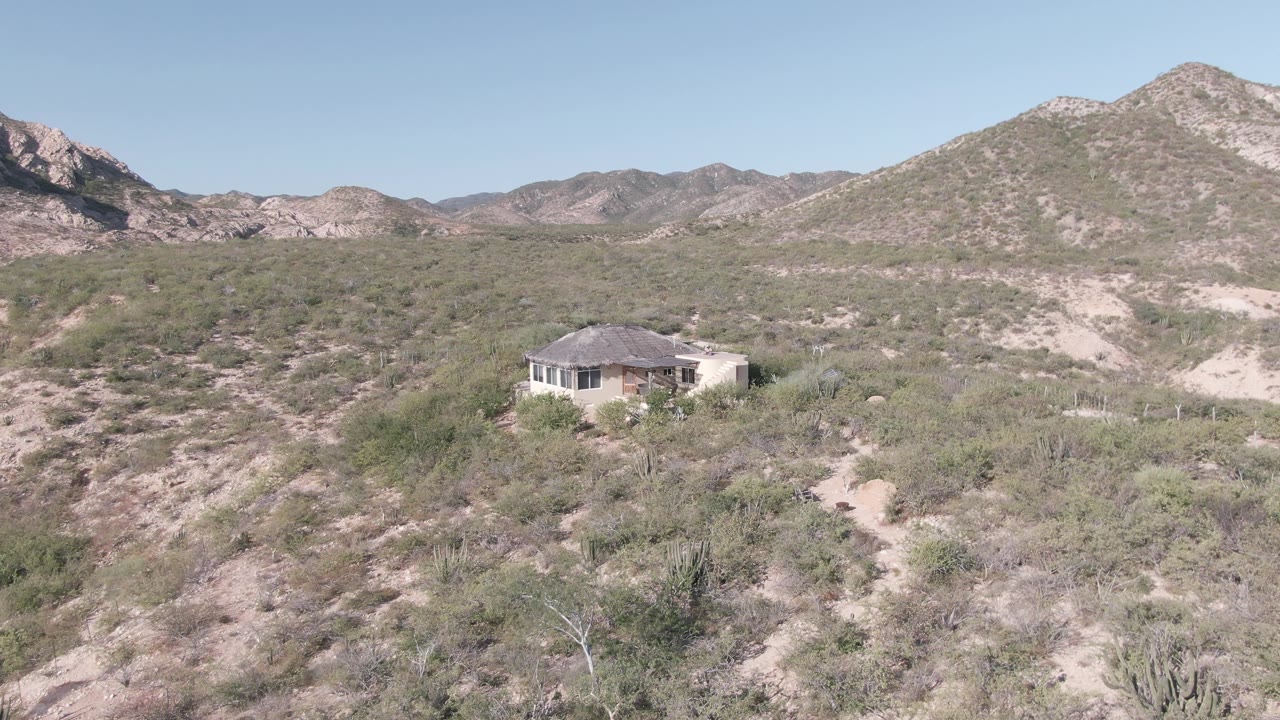 Elevated Cabo Pulmo desert frame showing the small light-colored house slightly closer, surrounded by scrubby vegetation, dry slopes, distant mountains, and no visible people.