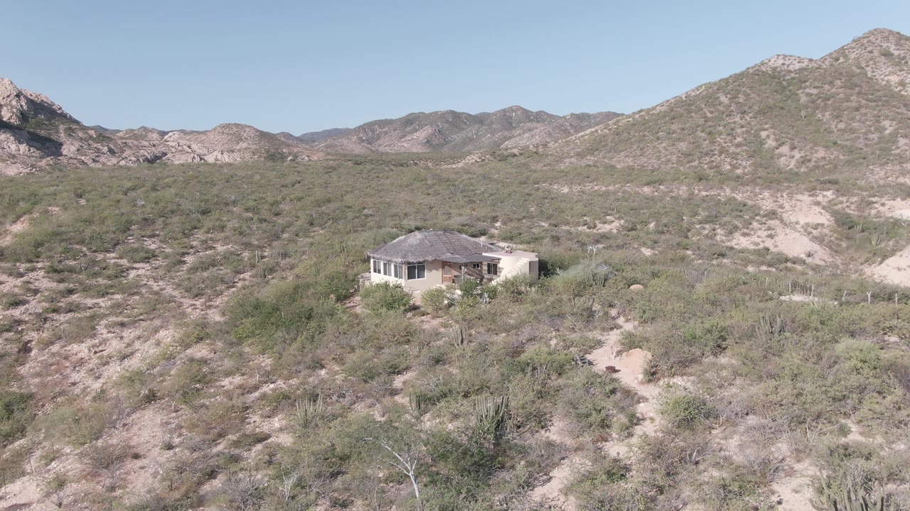 Aerial-looking view in Cabo Pulmo with a small house set amid dry brush, faint sandy tracks, rounded mountain slopes, pale blue sky, and no visible people.