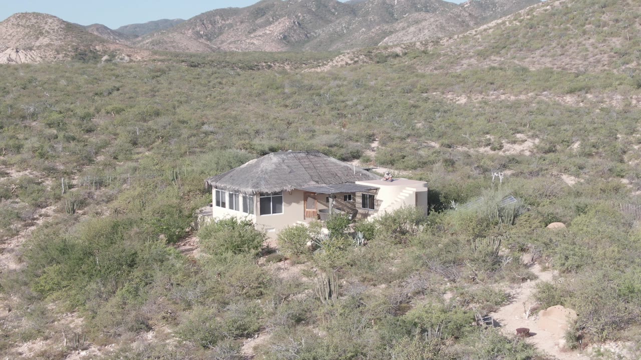 A small light-colored house sits in desert vegetation near Cabo Pulmo, Baja California Sur, with rocky mountains behind it and no visible people.