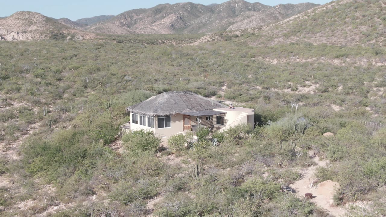 The house appears slightly farther into the frame within Cabo Pulmo desert scrub, with pale tracks, cactus shapes, and distant mountains visible, and no visible people.