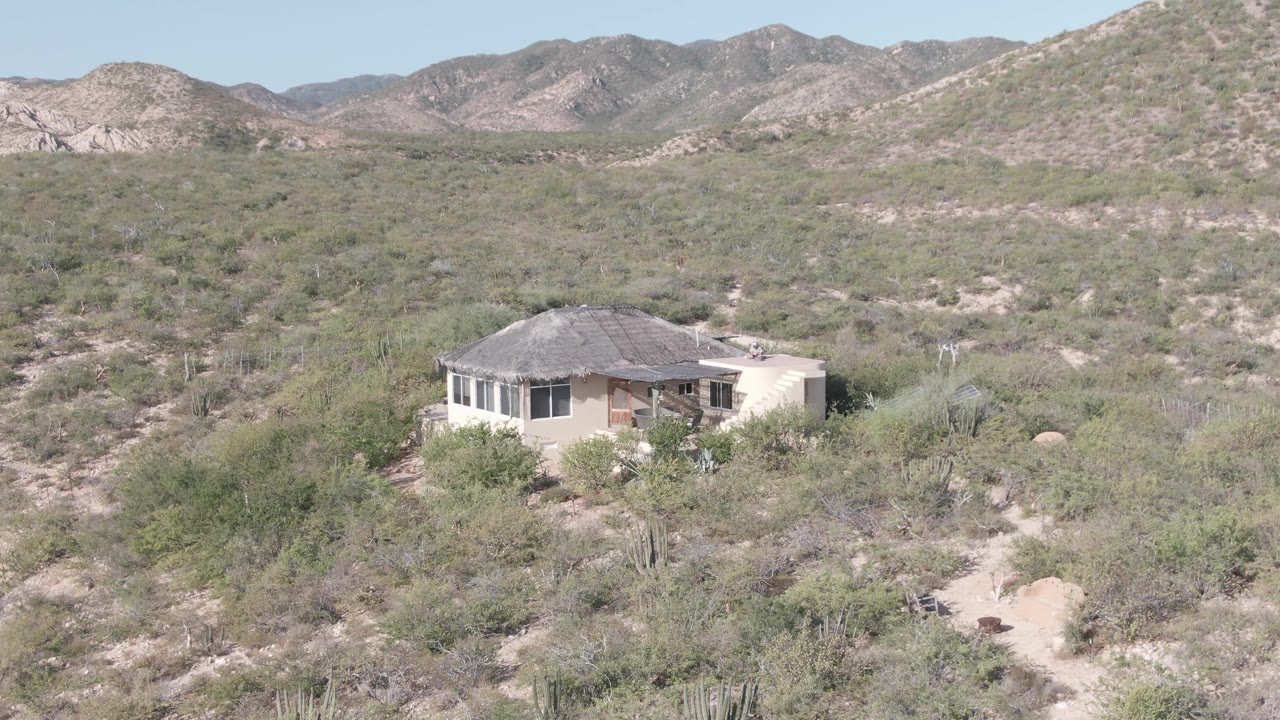 A small house remains in the lower-left desert foreground near Cabo Pulmo, with green-gray brush across the valley floor, a sloped hillside at right, and no visible people.