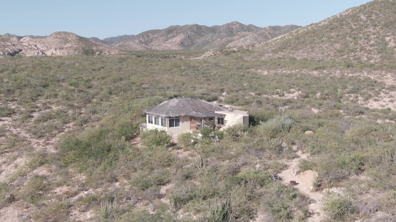 Elevated view in Cabo Pulmo, Baja California Sur showing a small house set in desert shrubs and cacti, with rocky mountains under a clear sky and no visible people.
