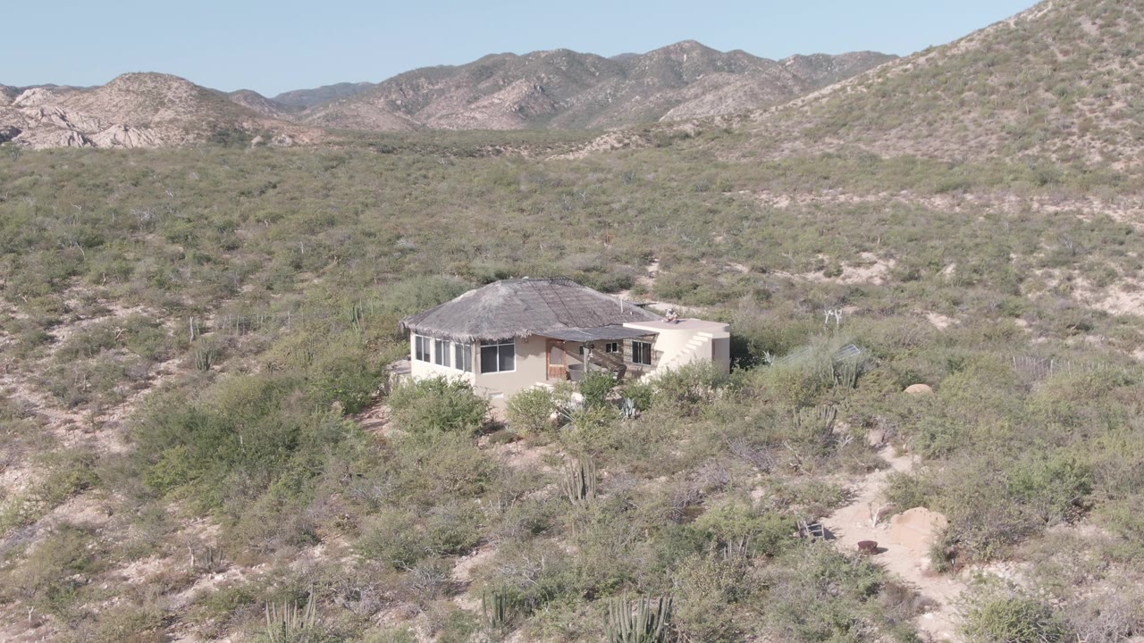 Cabo Pulmo aerial-style frame with a small single-story house surrounded by green-gray desert vegetation, pale sand, distant mountains, and no visible people.