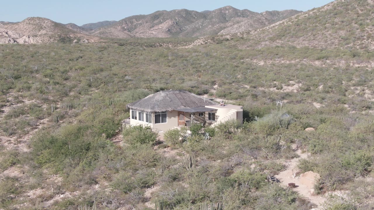 Elevated Cabo Pulmo view of a desert residence partly surrounded by shrubs and cacti, with layered rocky hills in the background and no visible people.