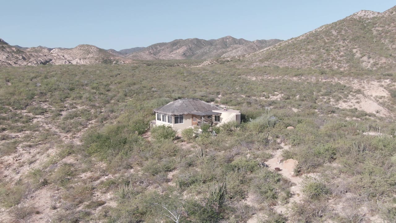 A small house stands in a scrubby Cabo Pulmo desert landscape below rocky Baja California Sur hills, with sandy ground and no visible people.