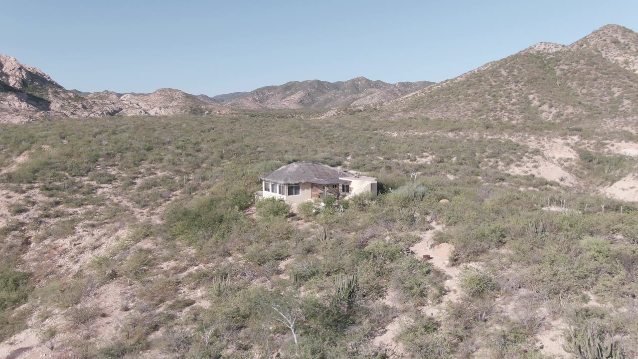 A light-colored house sits low in the Cabo Pulmo desert, with cactus and scrub across the foreground and layered rocky hills in the distance, with no visible people.
