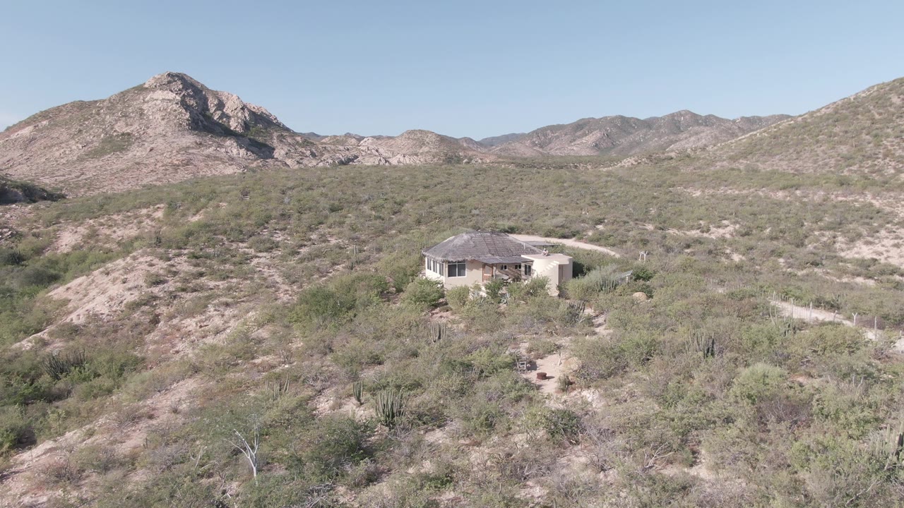 Aerial frame from Cabo Pulmo, Baja California Sur showing a light-colored house beside a pale dirt path, dry desert vegetation, and rocky mountains under a clear sky.