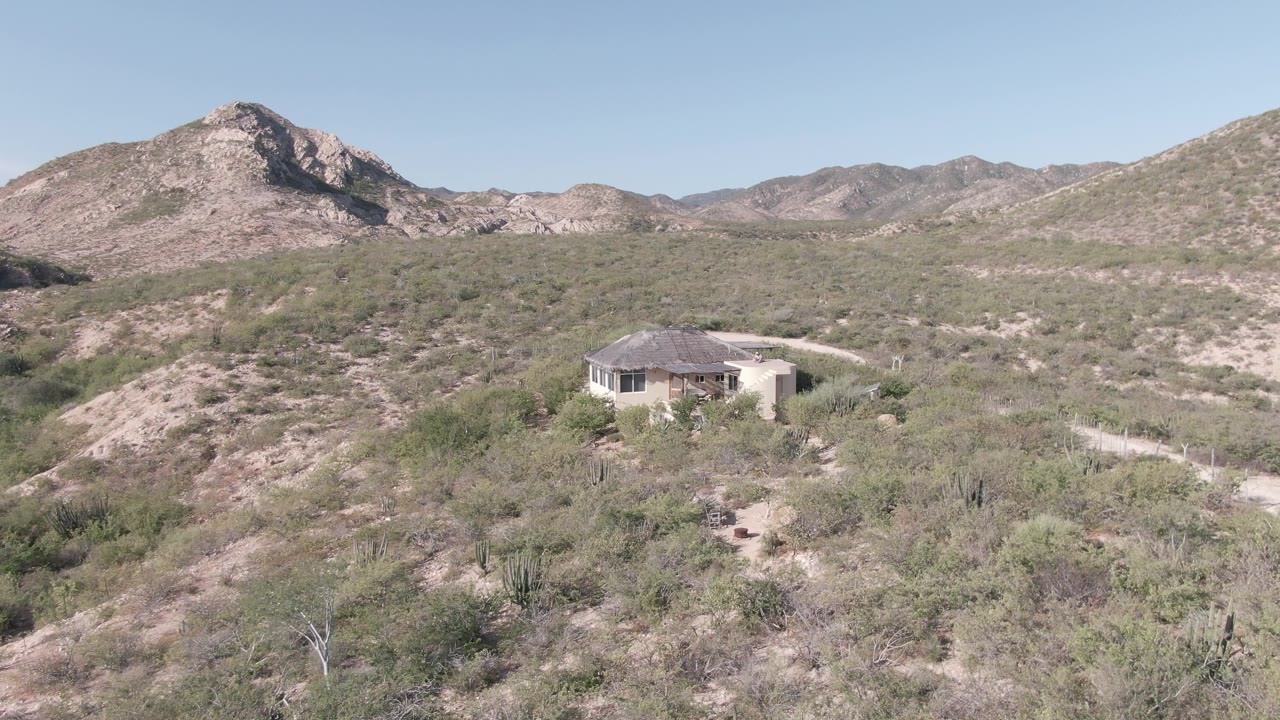 Desert hillside with a small house, winding path, scrub, and mountain ridge.