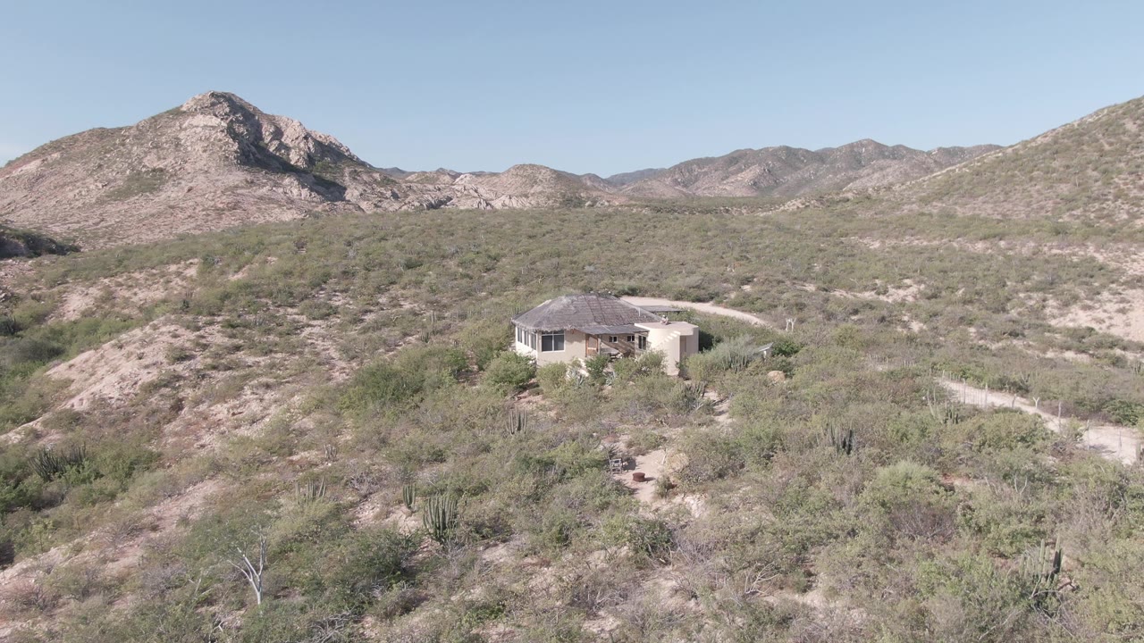 Cabo Pulmo aerial frame showing the house in the midground, pale footpaths through dense dry vegetation, and layered rocky mountains beneath a cloudless blue sky.