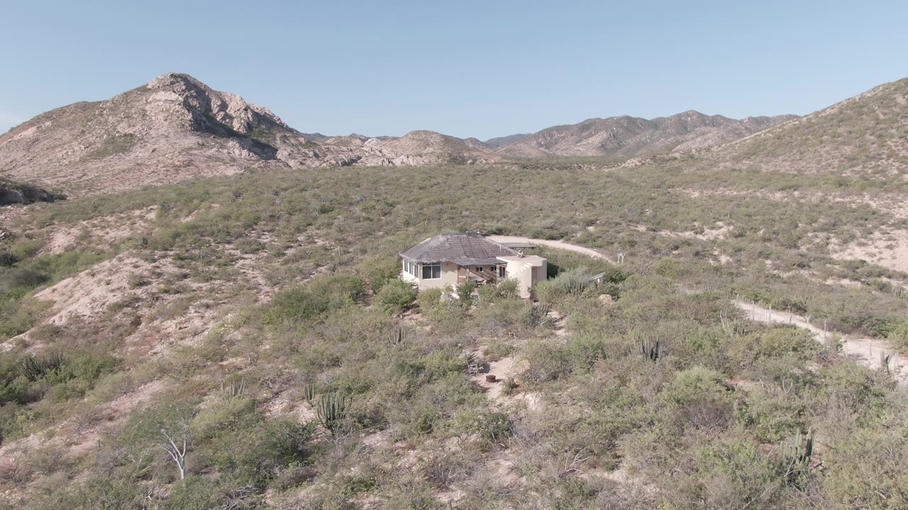 Aerial desert landscape with house, dirt path, scrubland, and rocky peak.