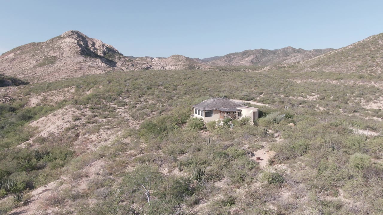 Desert hillside house framed below rocky mountains and pale sky.