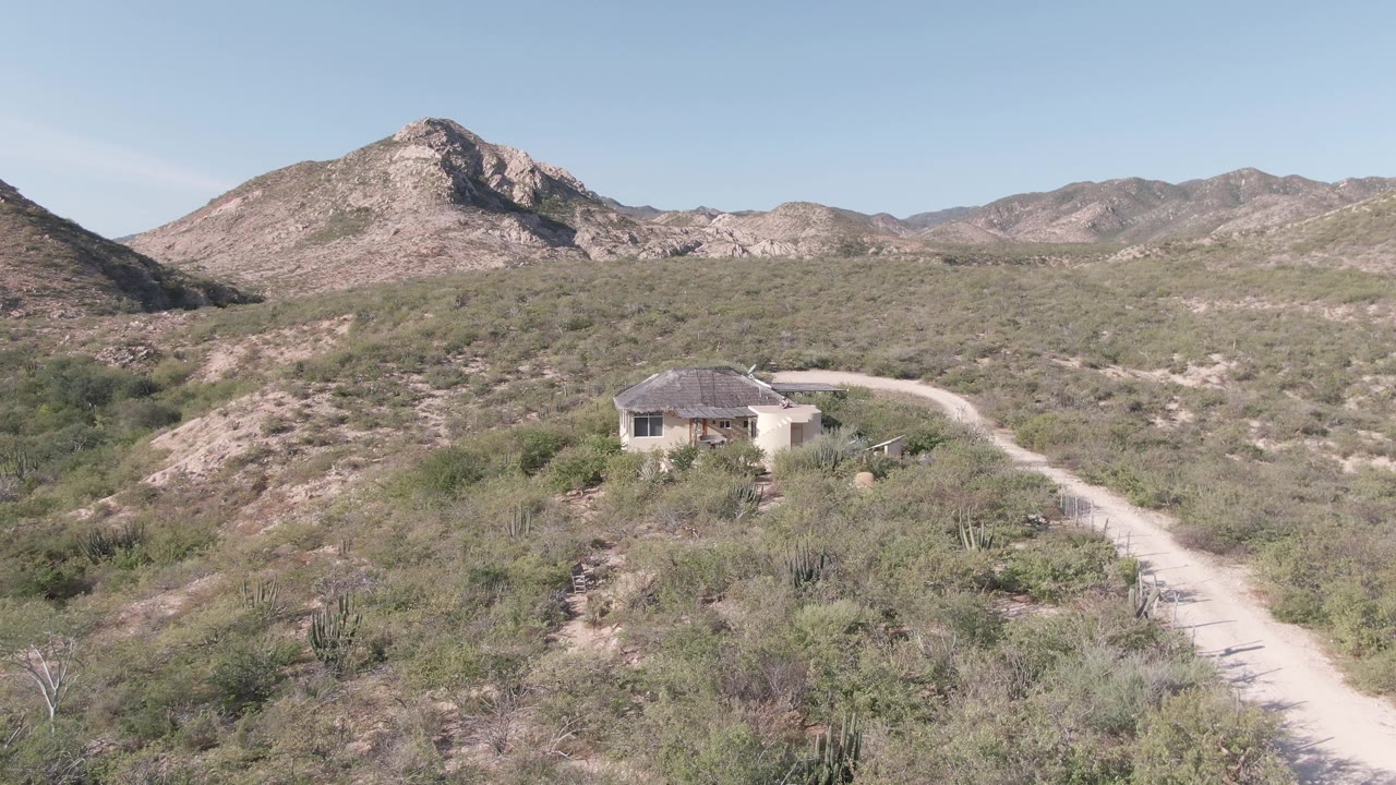 Cabo Pulmo desert frame with the road curving from the lower right toward a small building, low brush across the foreground, rocky mountains behind, and no visible people.