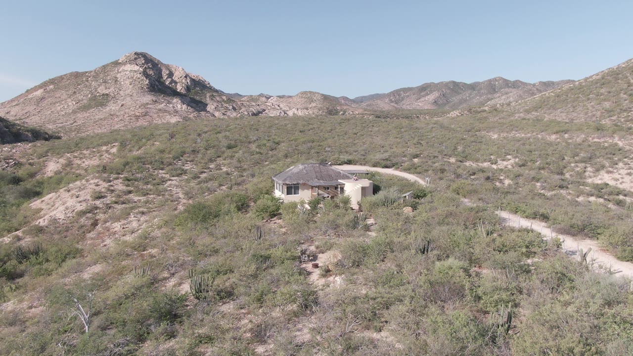 Cabo Pulmo desert hillside with a single-story house, pale dirt road, scrub vegetation, rocky mountains, and a small distant person near the house.