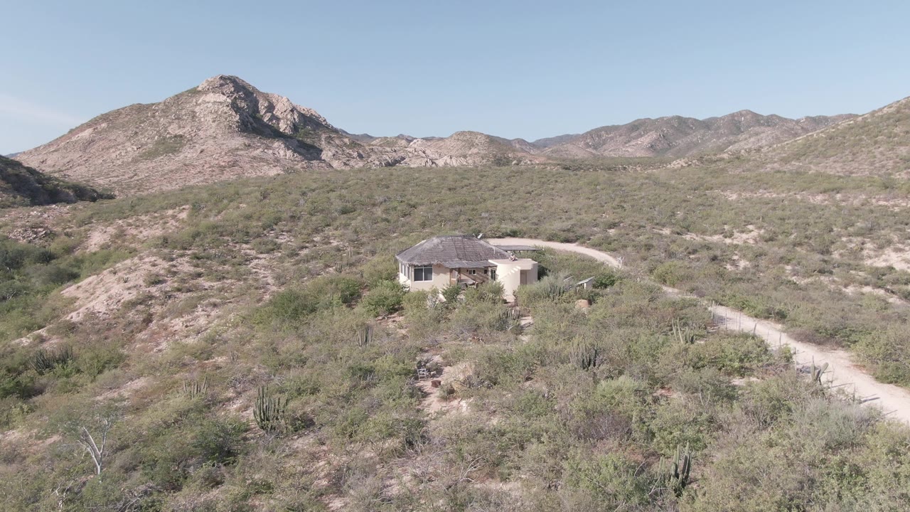 Cabo Pulmo desert landscape with a low house beside a pale dirt road, cactus and brush across the foreground, layered mountains in back, and no visible people.