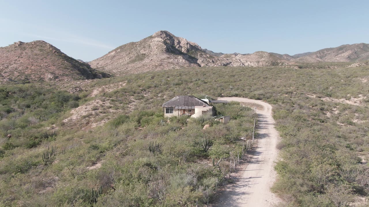 Early frame showing the pale road and small round-roofed building in Cabo Pulmo desert terrain, with green shrubs and no visible people.