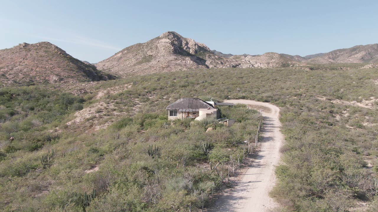 Mid-early frame with the dirt track bending through Cabo Pulmo shrubs toward the small structure, backed by sunlit rocky ridges and no visible people.