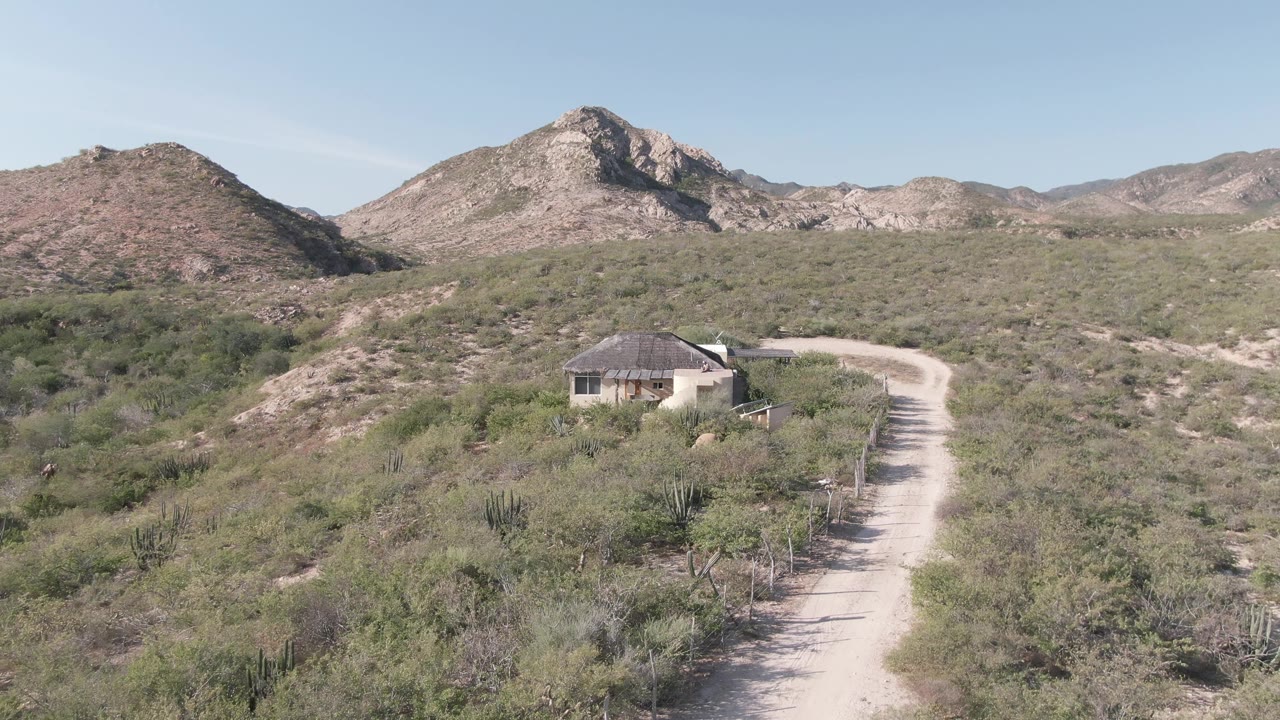 Midpoint frame placing the small building lower in the Cabo Pulmo desert scene, with scrub vegetation around the road and no visible people.