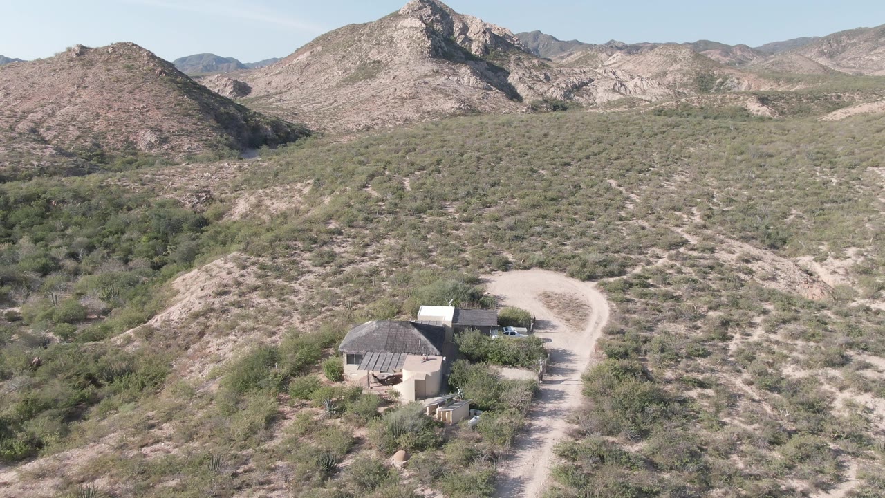 Aerial frame over Cabo Pulmo desert terrain with a dome-roof structure, a nearby roofed building, a pale dirt road, scrub-covered hills, distant rocky ridges, and no visible people.