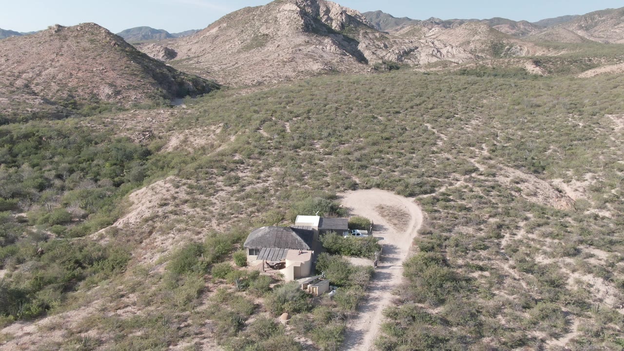 Wide Cabo Pulmo aerial with the dirt road curving past small desert structures in the lower frame, scrub vegetation filling the slopes, rocky mountains in back, and no visible people.