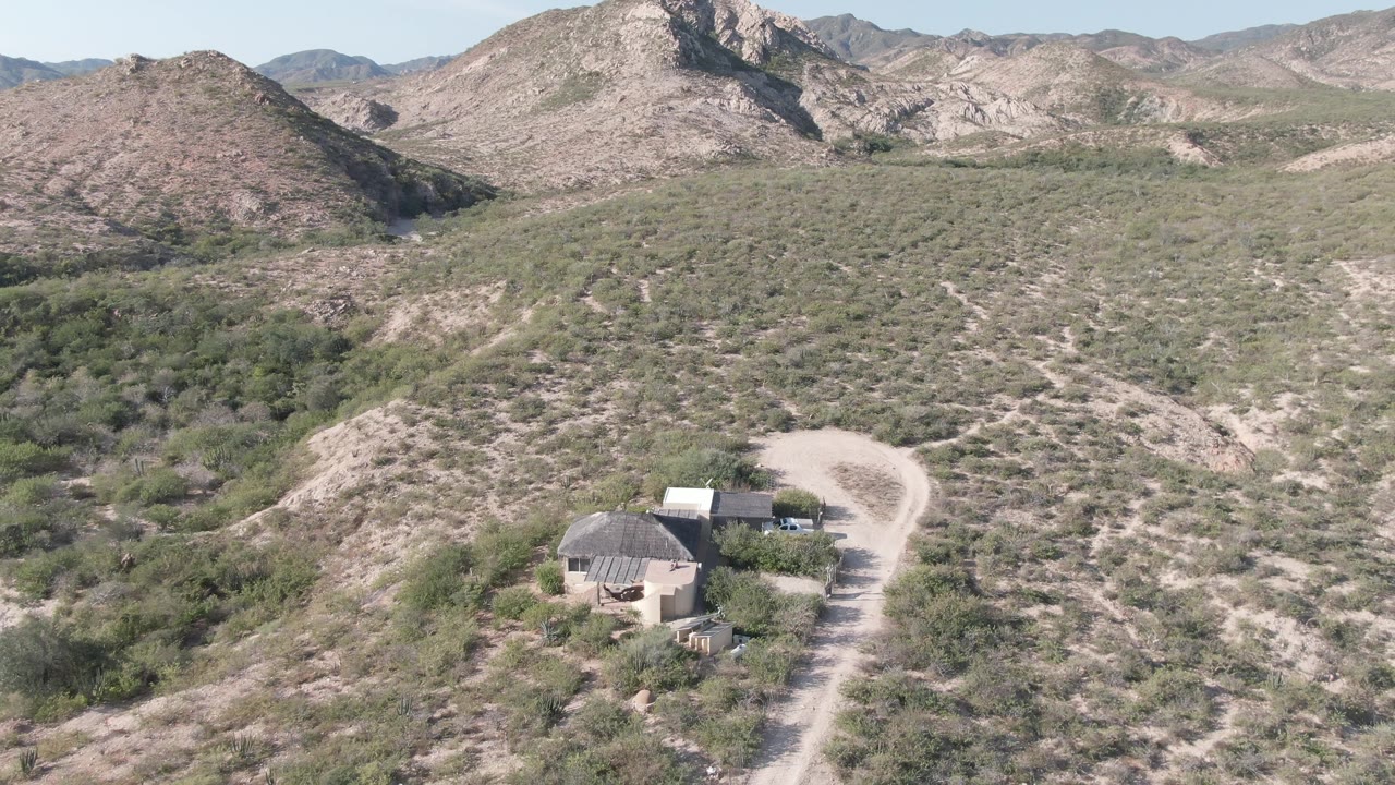 Cabo Pulmo drone view with the winding road brighter on the right side of the small structures, green-brown shrubs across the foreground slopes, layered rocky hills, and no visible people.