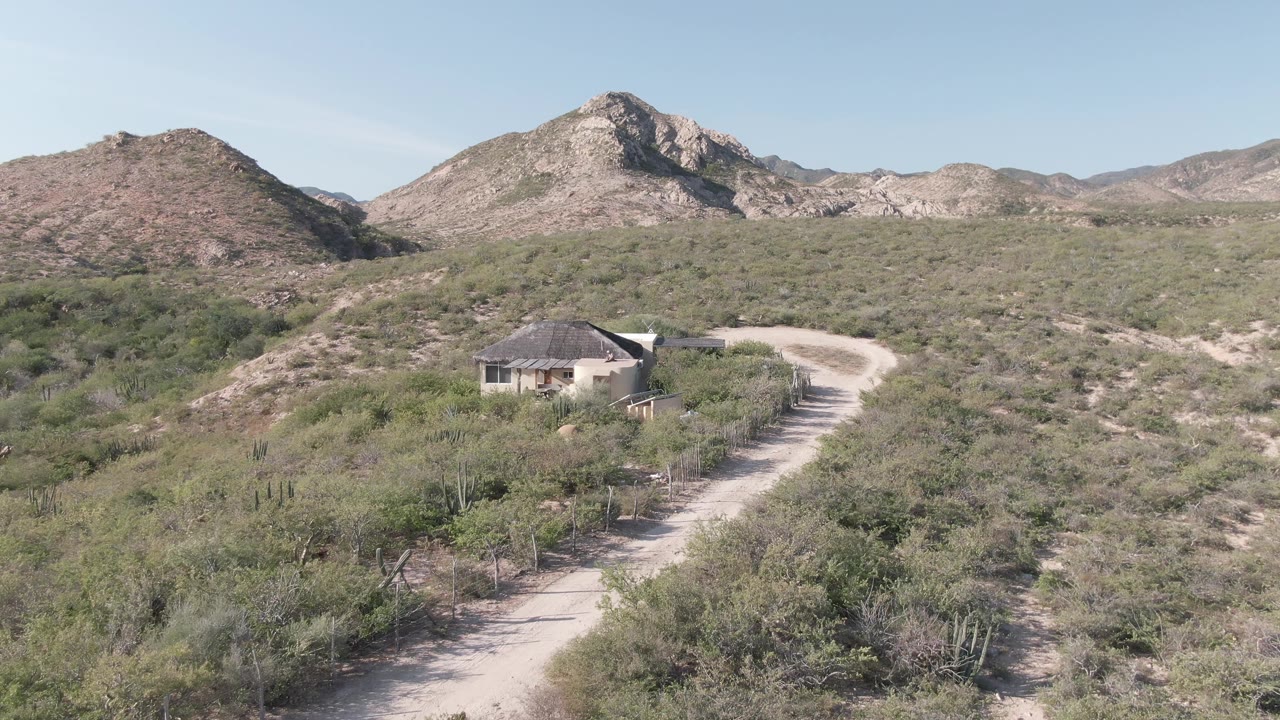 Opening frame of a small house on a scrubby hill in Cabo Pulmo, Baja California Sur, with a pale dirt path curving past cacti and no visible people.