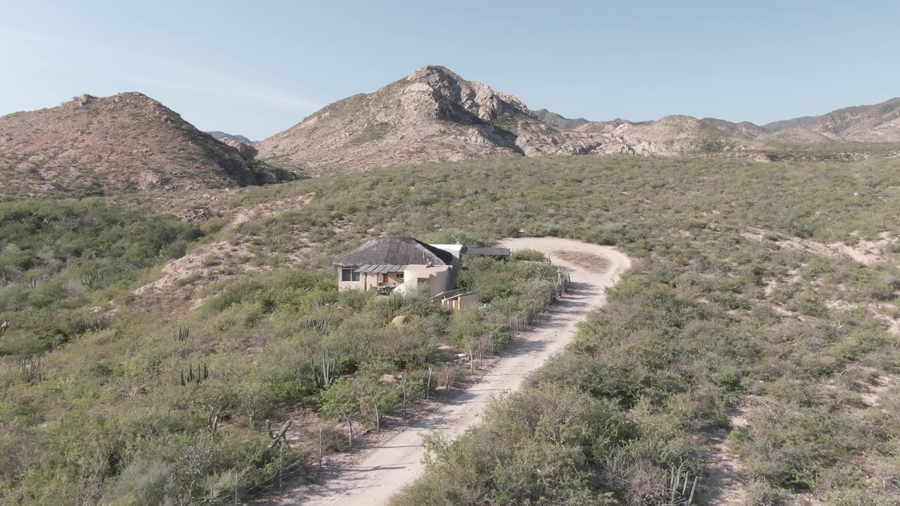 Aerial view of a small hilltop house, desert brush, dirt road, and mountains.