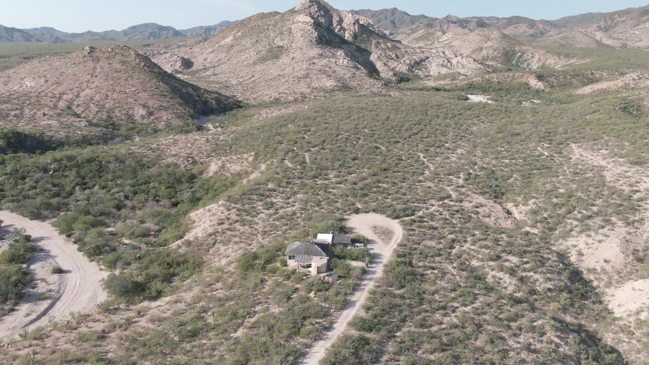 Aerial frame in Cabo Pulmo, Baja California Sur, with the house near the lower center, curving dirt roads on both sides, dry shrubs, and no visible people.