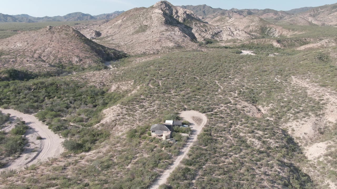 Wide aerial view over Cabo Pulmo desert terrain, with a small house near the lower right, scrub-covered slopes, layered rocky mountains, and no visible people.