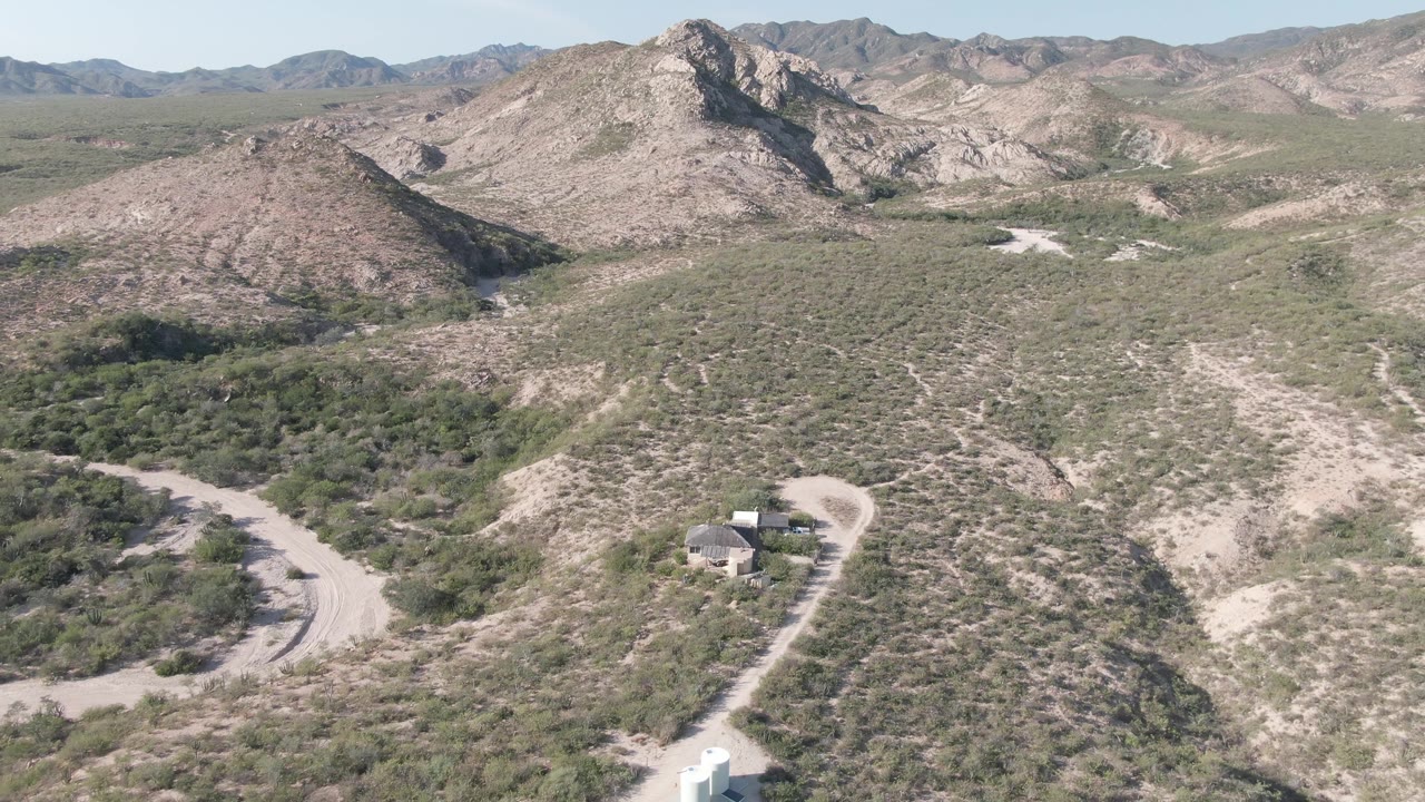 Wide aerial frame over Cabo Pulmo, Baja California Sur, with a white vehicle near the lower road, small roadside structures, scrub-covered hills, rocky mountains, and no visible people.