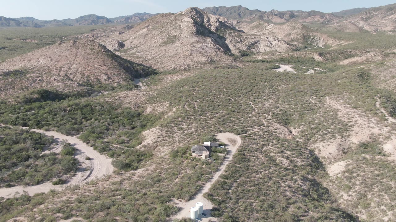 Aerial frame in Cabo Pulmo showing the white vehicle farther along the curving dirt road, with tan wash lines, green scrub patches, distant rocky ridges, and no visible people.