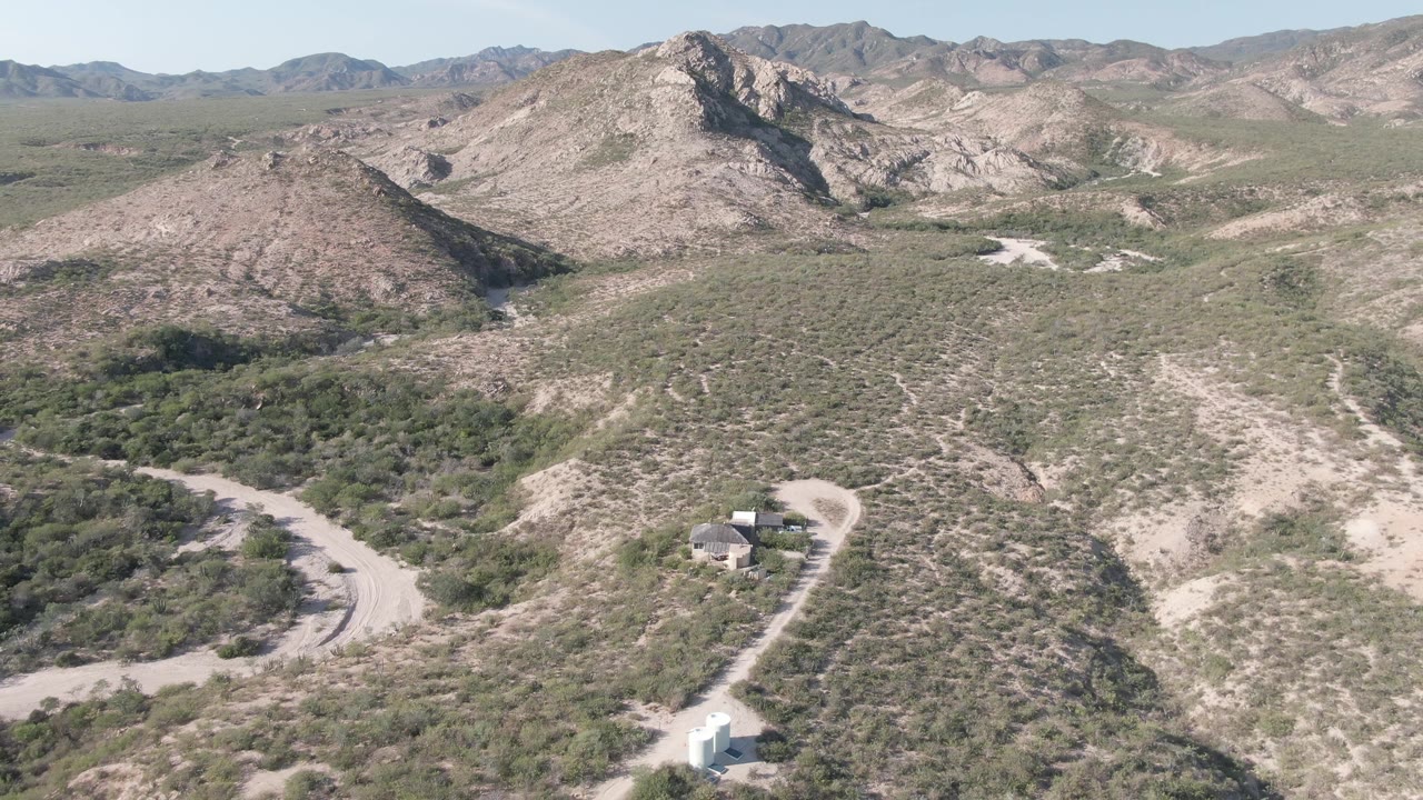 Wide Cabo Pulmo aerial view with the white vehicle and small light structures near the lower road, scrubby slopes filling the midground, rocky mountains across the horizon, and no visible people.