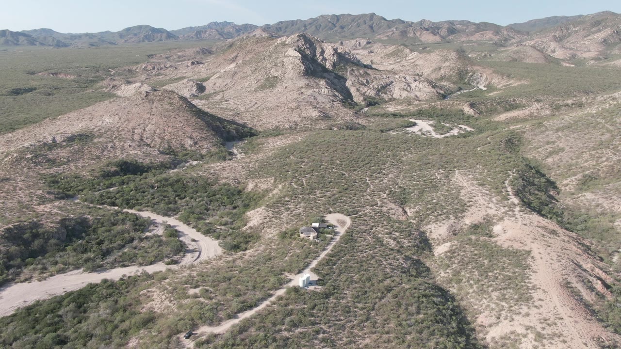Aerial frame over Cabo Pulmo, Baja California Sur, showing a winding pale dirt road, scrub-covered hills, distant ridges, and no visible people at the start of the clip.