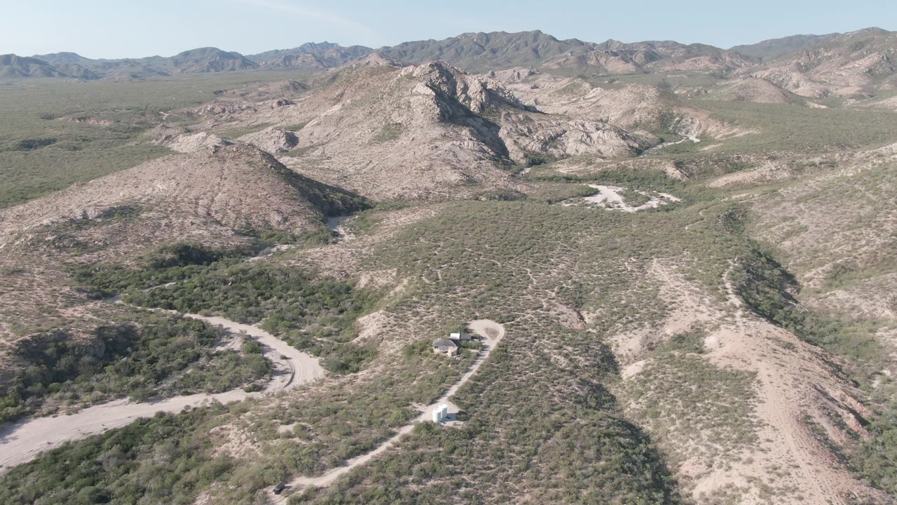 Aerial view in Cabo Pulmo with the dirt road curving across the lower hills, patchy green vegetation on rocky slopes, distant mountain ridges, and no visible people.