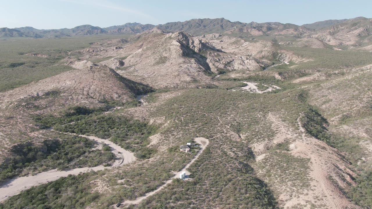 Cabo Pulmo aerial frame showing intersecting pale dirt tracks below rocky hills, low green brush, a long ridge line in the background, and no visible people.
