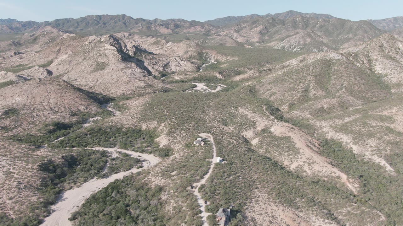 Aerial frame over Cabo Pulmo, Baja California Sur, showing a pale winding road, scrub-covered hills, distant ridgelines, and no visible people.