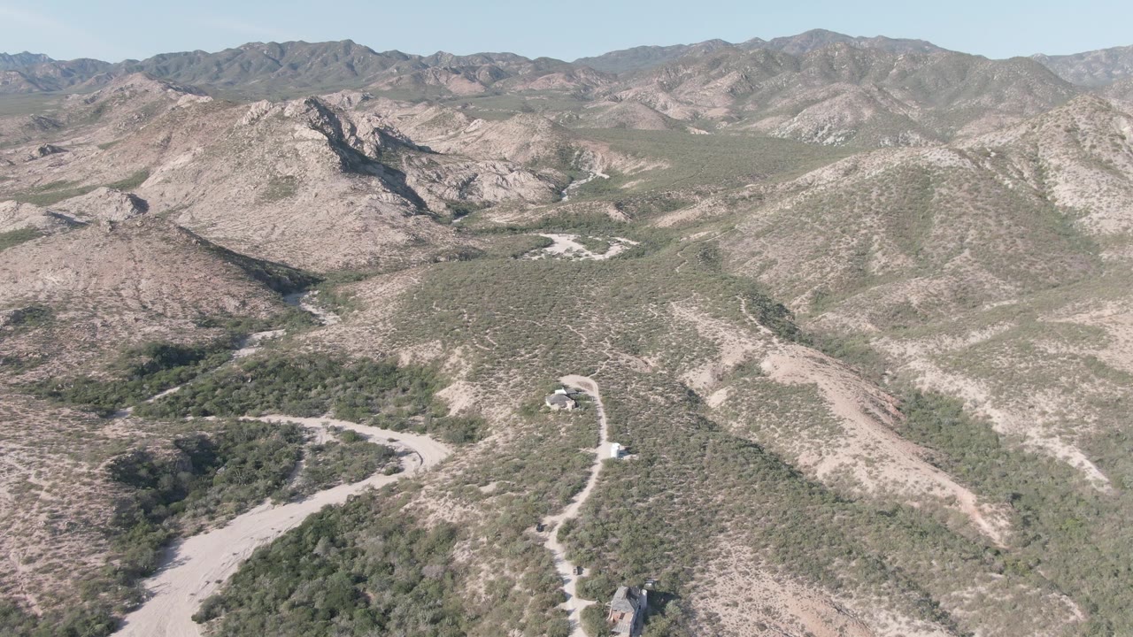 Aerial view in Cabo Pulmo with the curving road slightly closer, scrub vegetation lining the wash, rocky slopes behind it, and no visible people.