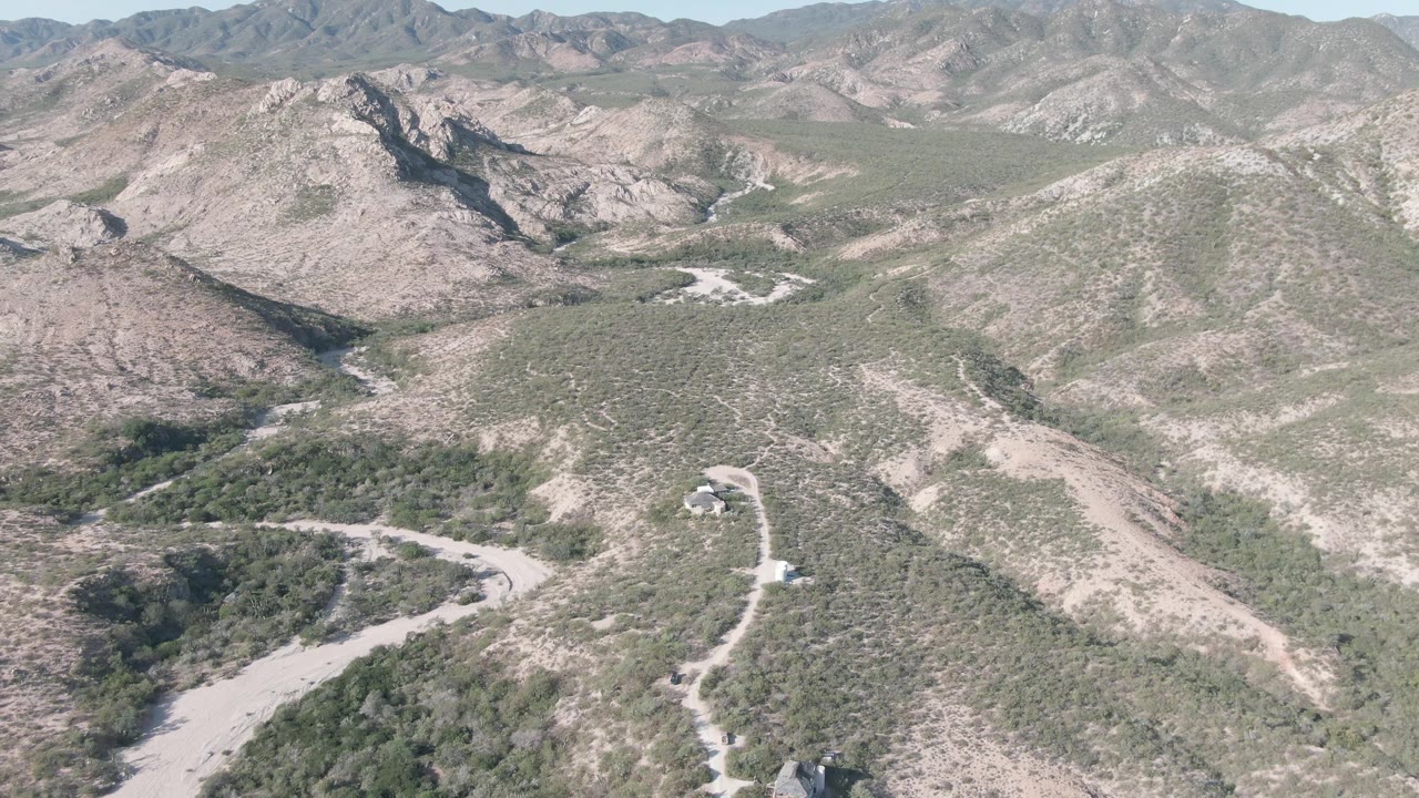 Aerial frame in Cabo Pulmo showing the winding road more centrally through tan rocky slopes, green brush patches, distant mountains, and no visible people.
