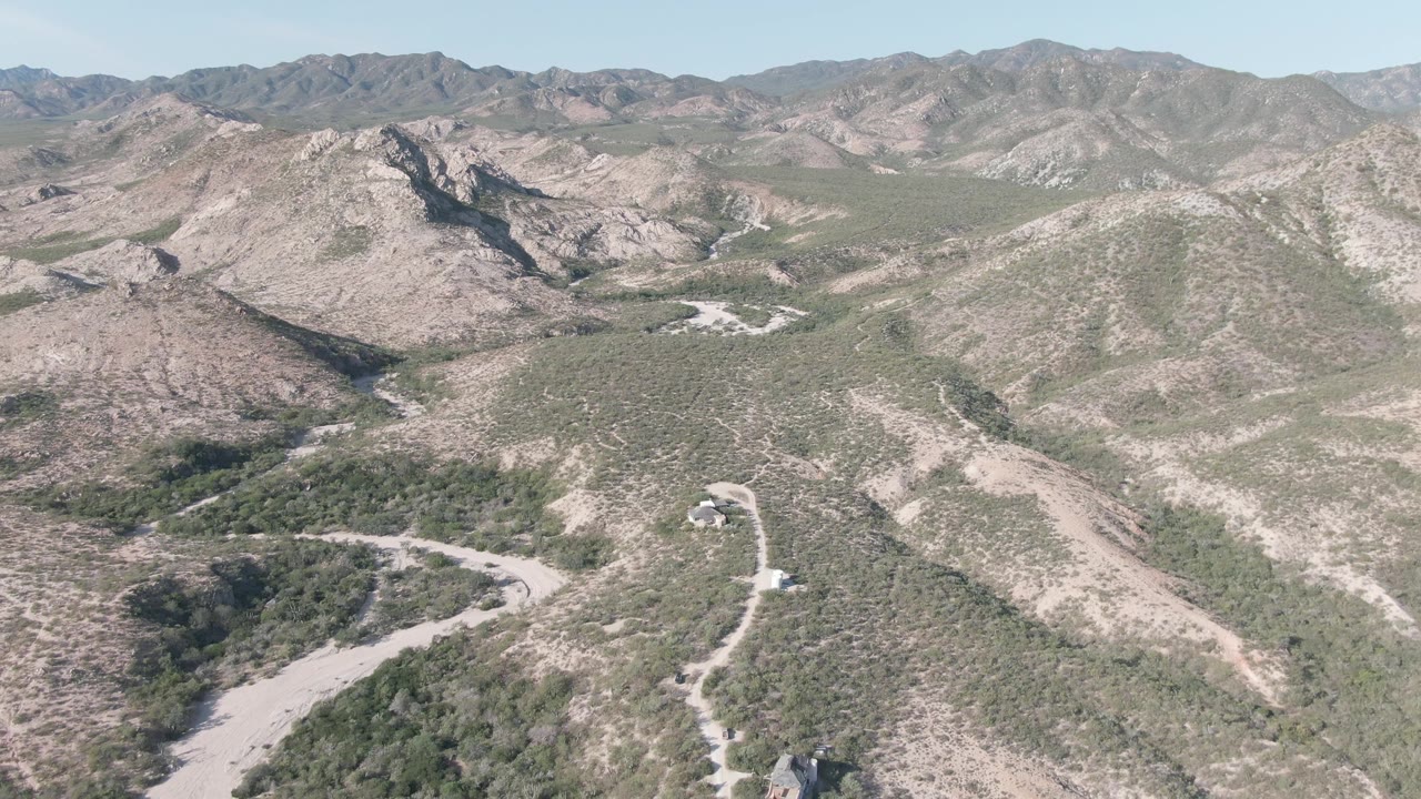Mid-clip aerial view over Cabo Pulmo's rocky hills, with a pale road crossing the lower frame, scrub vegetation on slopes, layered mountain ridges, and no visible people.