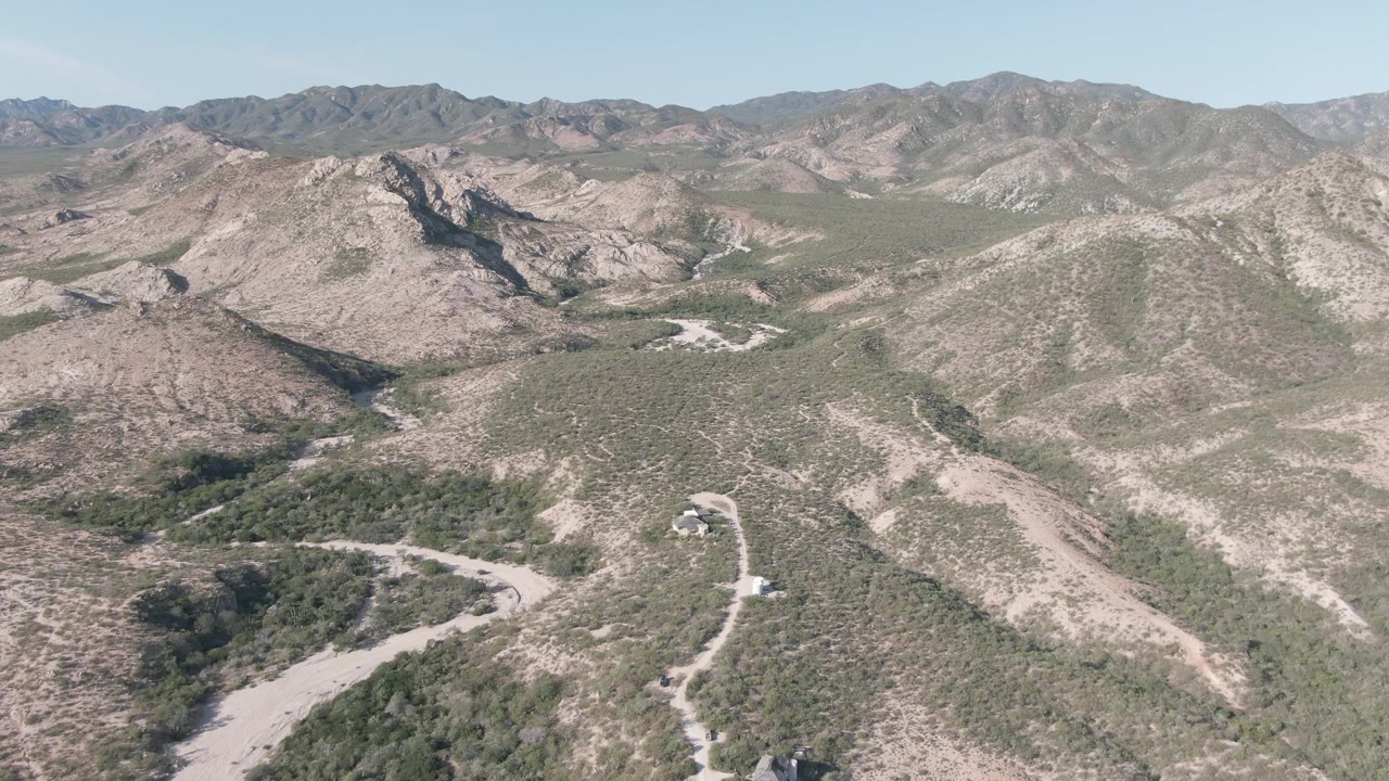 A later aerial frame over Cabo Pulmo with the road curving near the lower left, a larger sweep of rocky hills across the center, distant ridges, and no visible people.