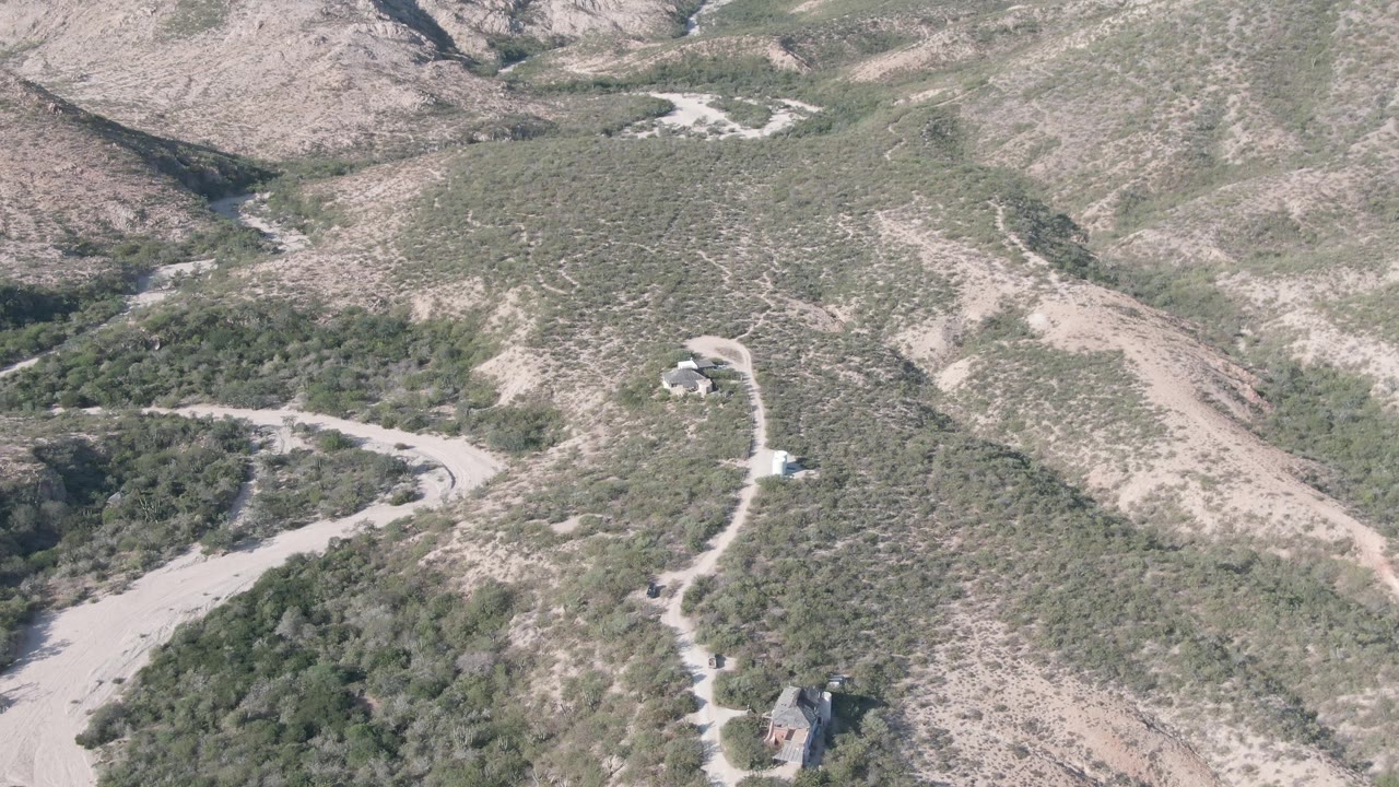Opening aerial frame over Cabo Pulmo, Baja California Sur, showing scrub-covered brown hills, a winding dirt road, a dry wash, and small structures with no visible people.